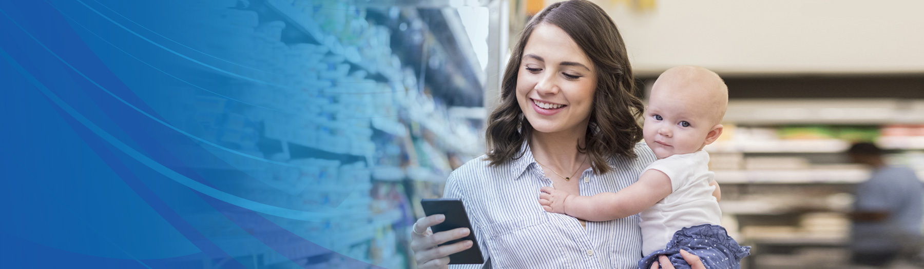 Young mother in the supermarket holding her baby, looking at a smartphone