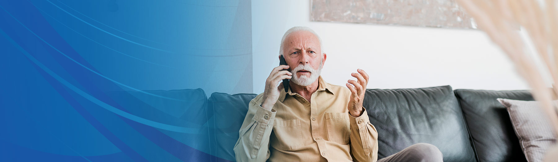 Older gentleman talks on a mobile phone and gestures while sitting on a couch at home.